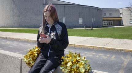 student holding phone and sitting in front of high school with cheerleading pom-poms beside her