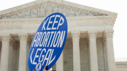 two hands holding round sign reading "Keep Abortion Legal" with Supreme Court building in background