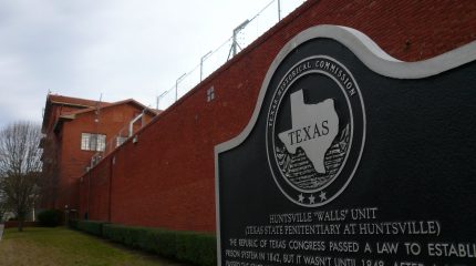 Texas historic marker in front of brick prison wall topped with barbed wire.