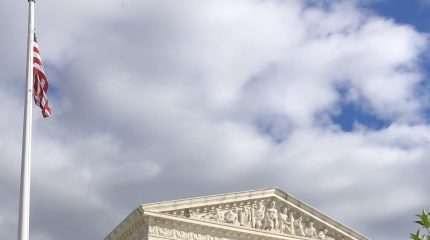 top of supreme court building with american flag on left against cloudy blue sky