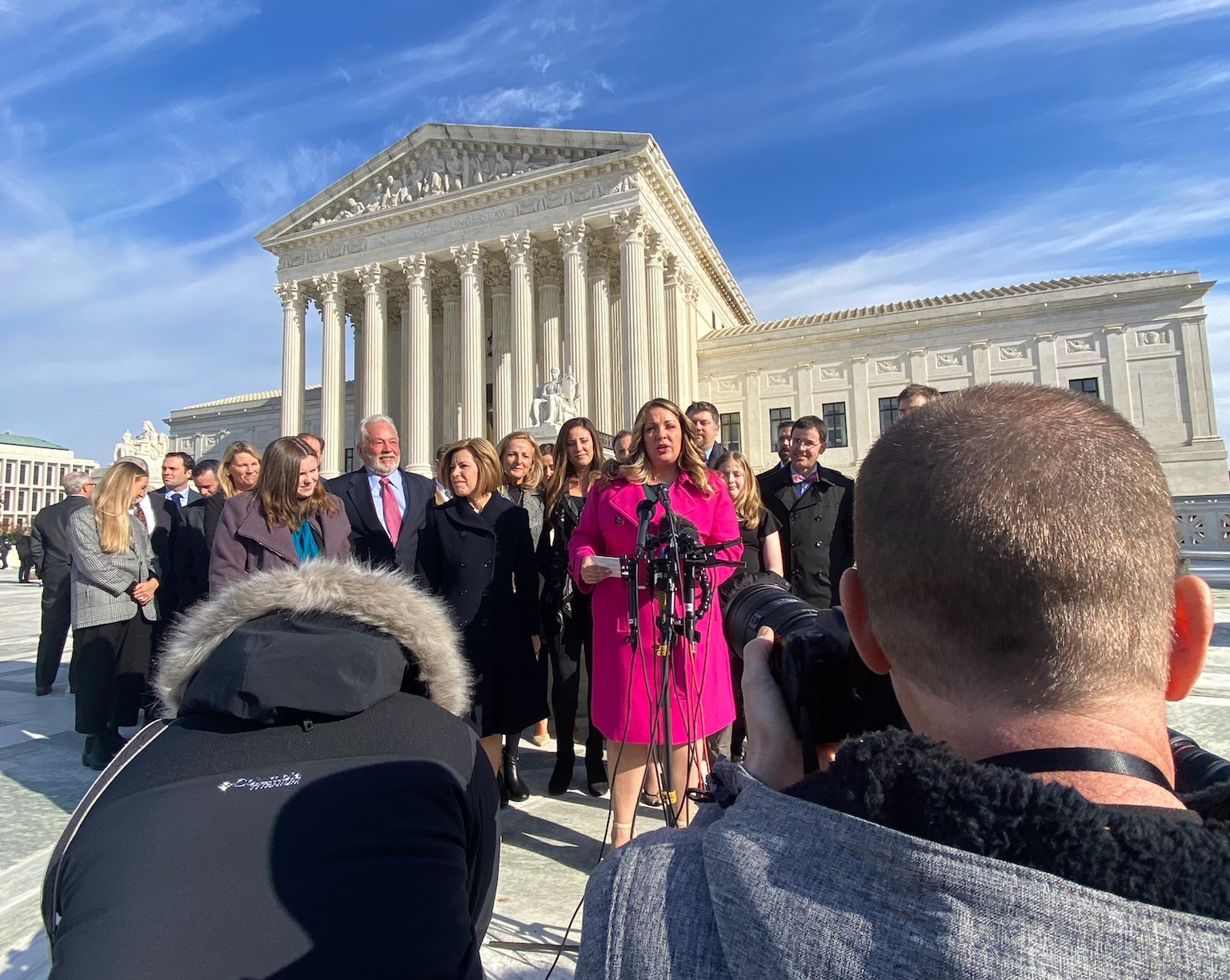 A woman in a winter coat speaks at a microphone, surrounded by supporters, with the Supreme Court building in the background.
