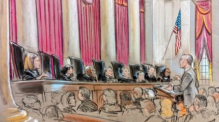 Eight justices on the bench speaking to and watching a man at the podium in a crowd of people.